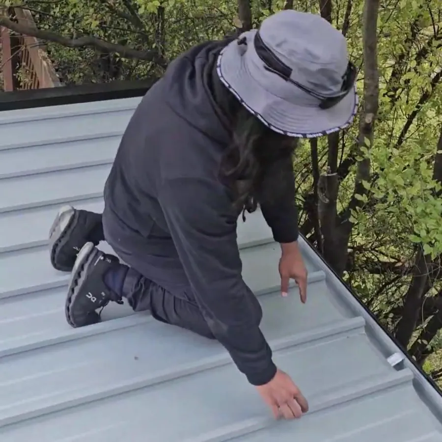 Roofer in a bucket hat kneeling on a new pale grey metal roof beside gum trees